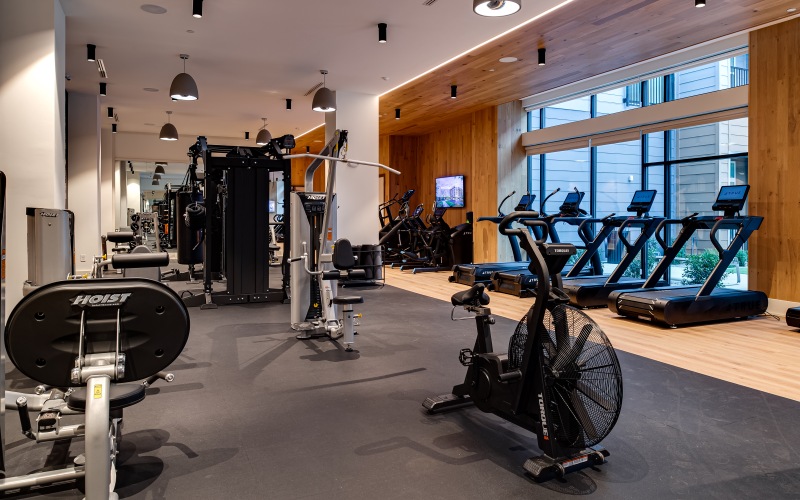 a well lit gym with treadmills bikes and stair stepper with a tv on the wall 
